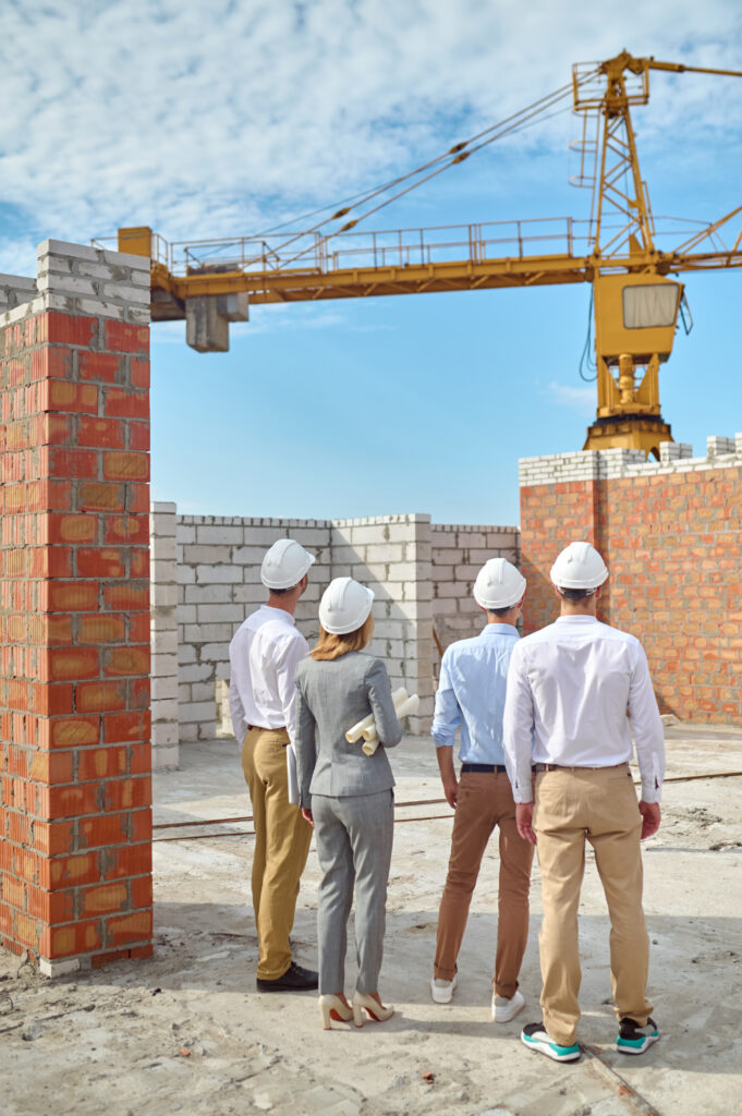 Back view of a supervisor with technical drawings and builders standing on the construction site